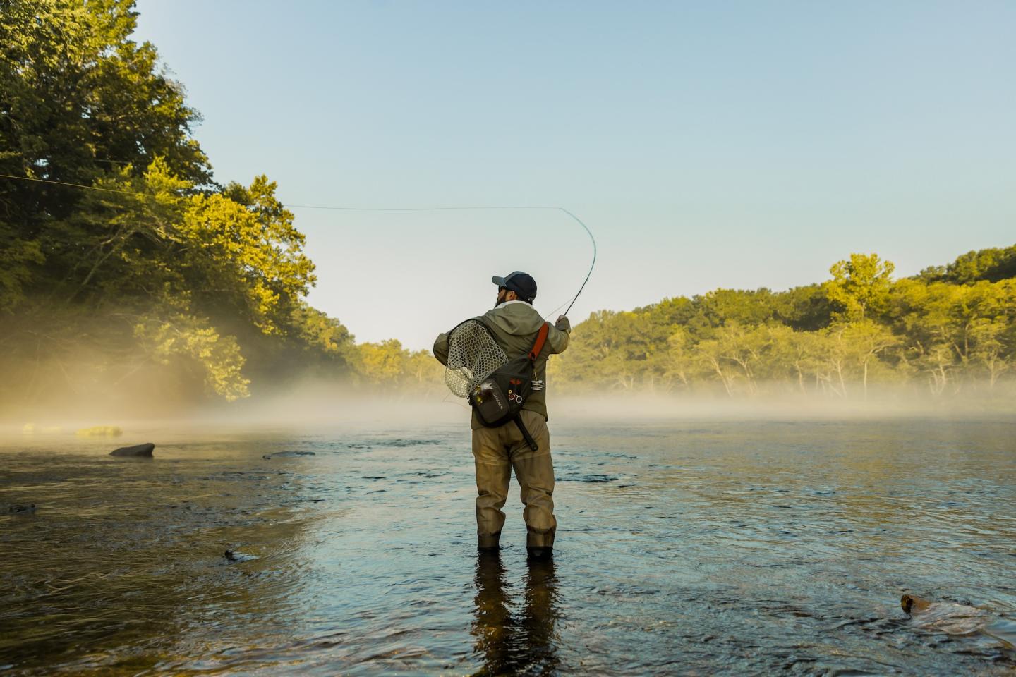 Angler fly-fishing in a tranquil, misty river at sunrise.