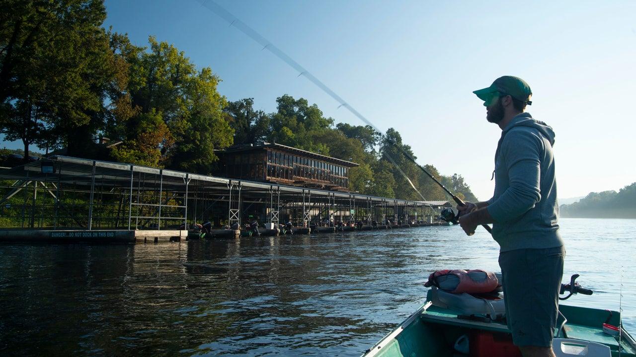 Man fishing from a boat near a dock, surrounded by trees and calm water.