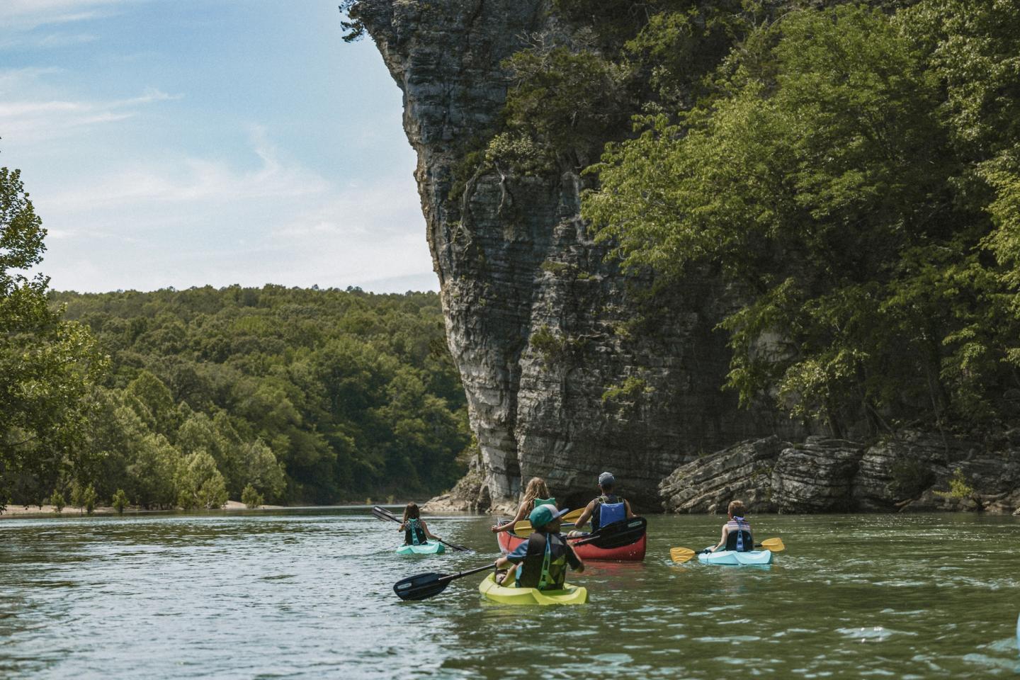 Kayakers paddle on a calm river near a rocky cliff with lush greenery.