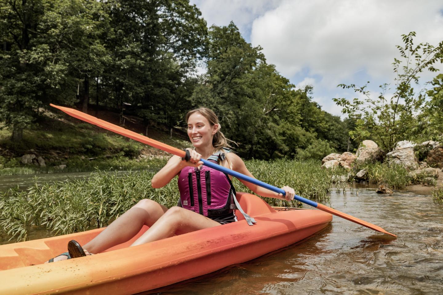 Young woman kayaking on a calm river, smiling under a partly cloudy sky.