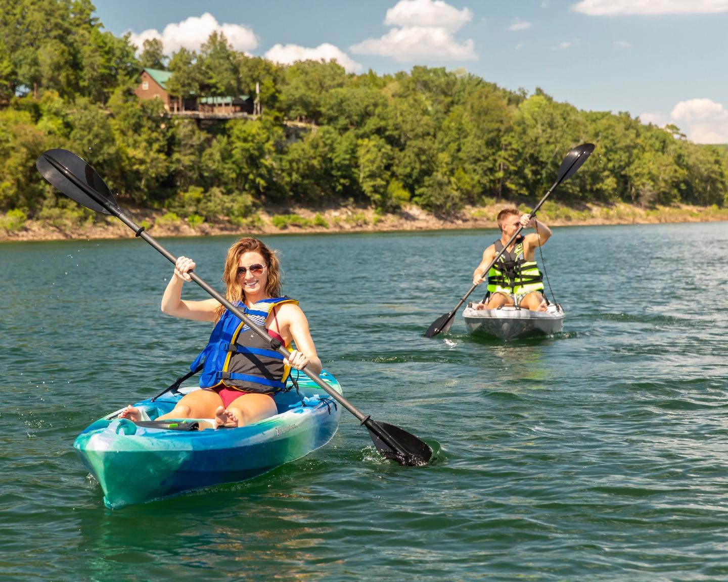 Two people kayaking on a lake with green trees and blue sky.