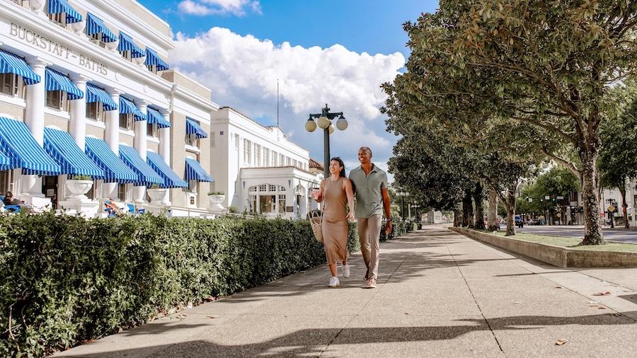 Couple walking on a sunny street lined with trees and a building with blue awnings.