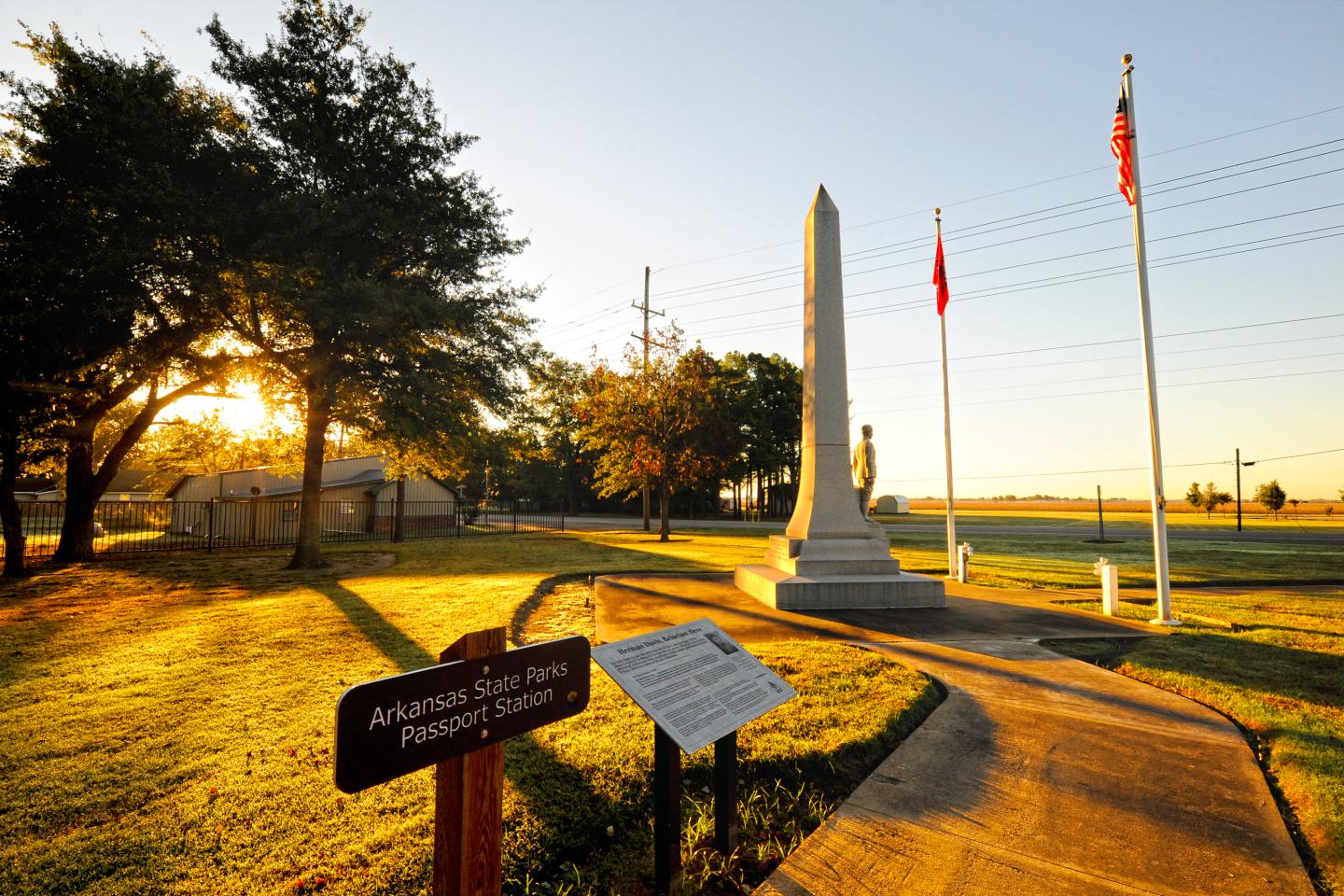 Sunlit park with an obelisk monument and flags, surrounded by trees and paths.