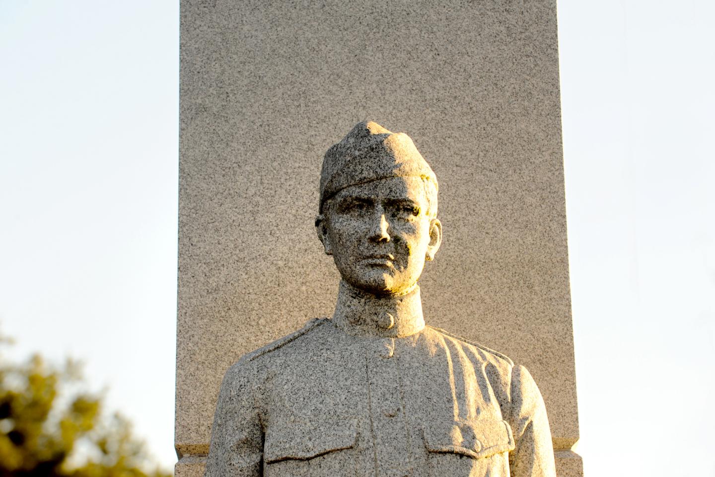 Stone statue of a uniformed soldier in front of a tall monument.