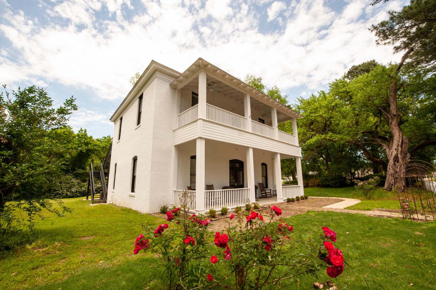 White two-story house with a porch, surrounded by green trees and red roses in the foreground.