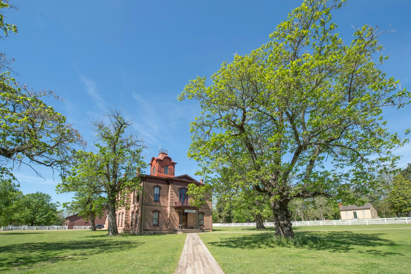 Historic brick building with a red roof, surrounded by green trees and grass, under a blue sky.