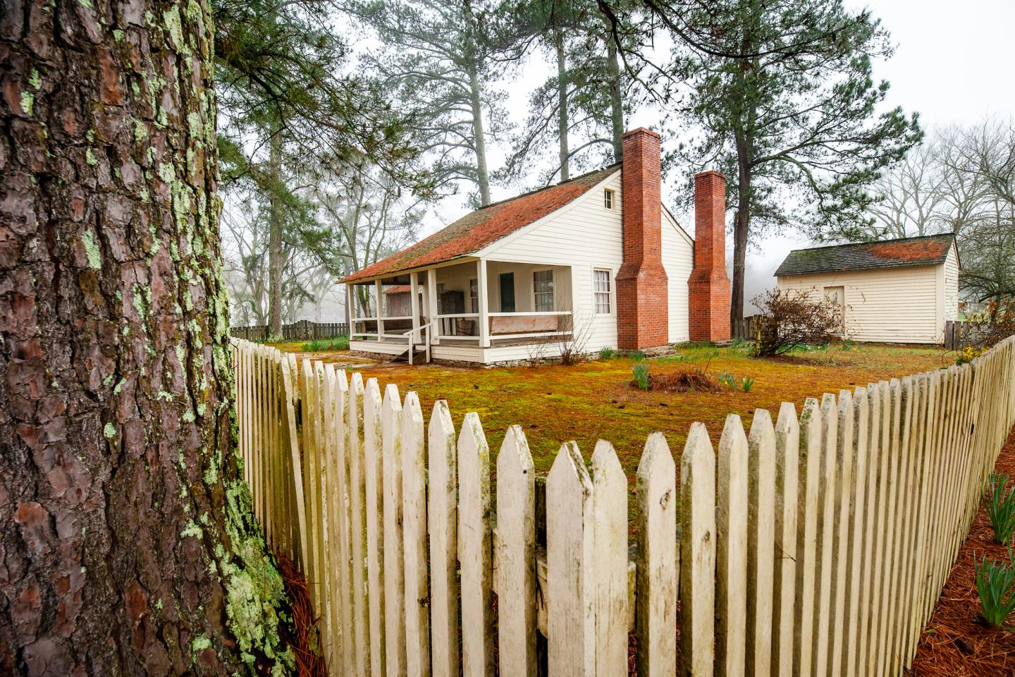 Cozy house with red chimneys behind a white picket fence, surrounded by tall trees.