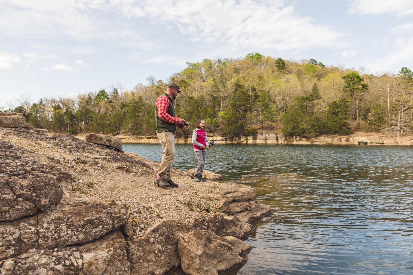 Father and daughter fishing on a rocky lakeshore under a cloudy sky.