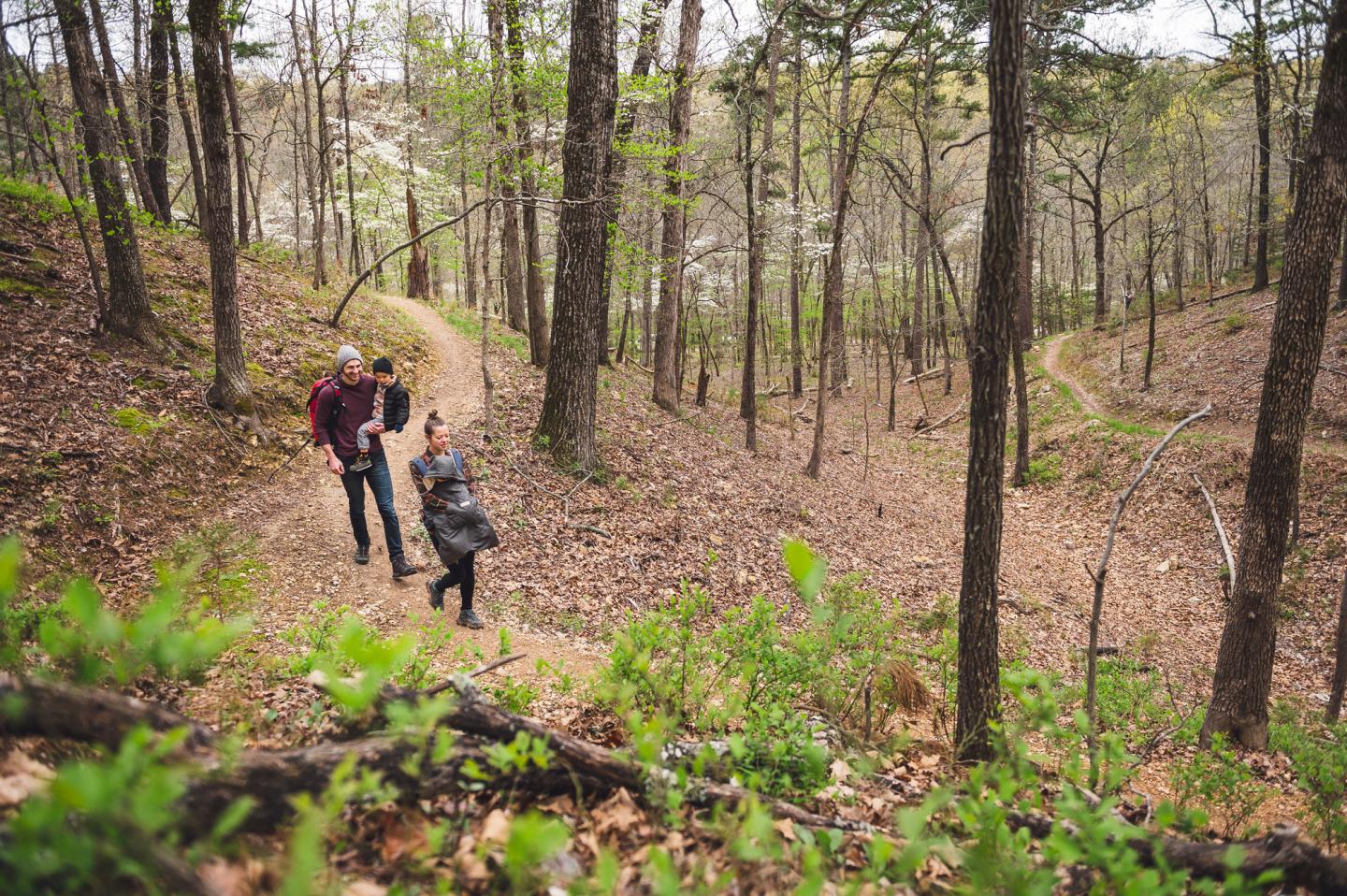 Two parents holding their children while hiking on a forest trail surrounded by trees.