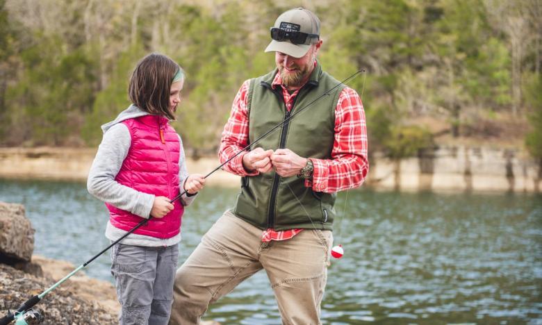 Man and child fishing by a lake, trees in background.