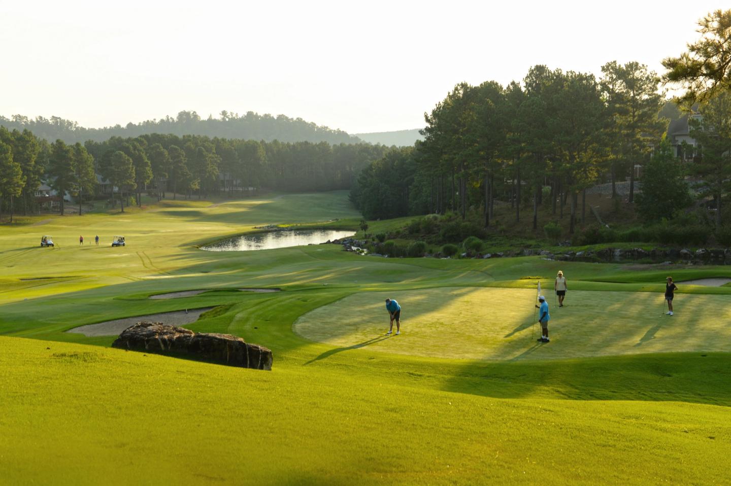 Golfers on a verdant course with trees and a pond in the background.