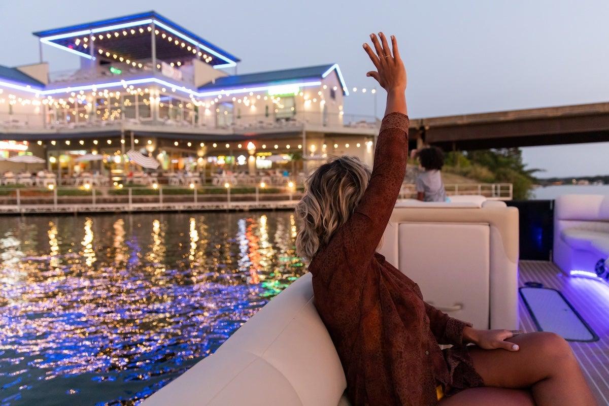 Woman on a boat raising hand, lit building on the water in the background.