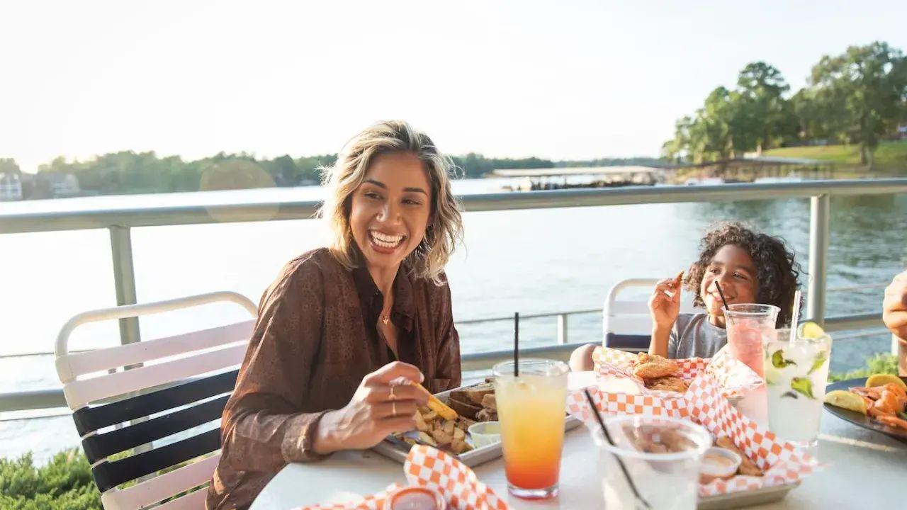 Two people smiling at a lakeside table with food and drinks.