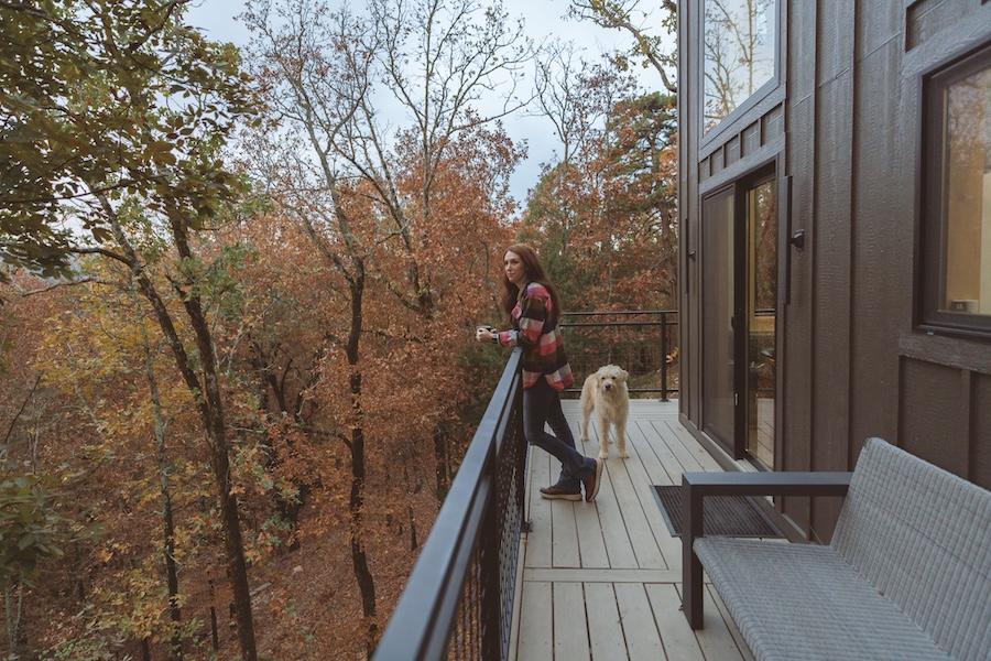 Woman and dog on cabin balcony with autumn trees in the background.
