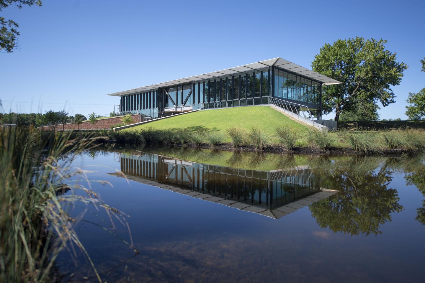 Modern glass building on a hill, reflected in a calm pond, under a clear blue sky.