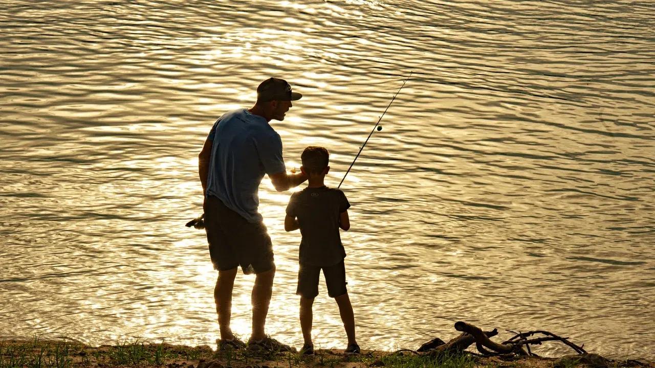 Silhouettes of man and child fishing at sunset by a lake.