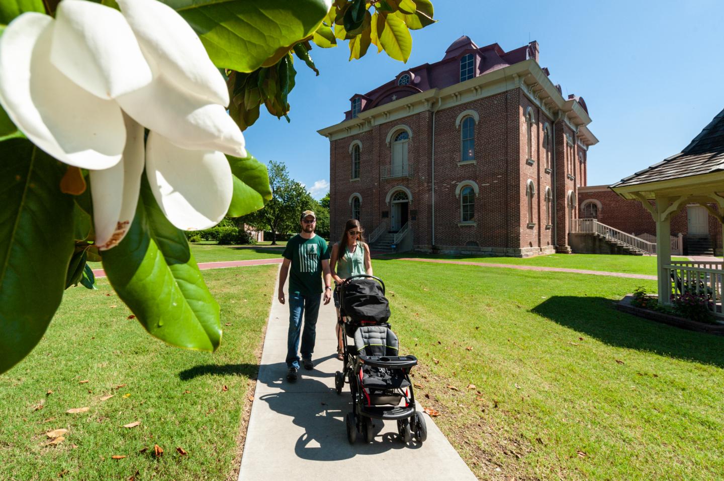 Couple with stroller walking on sunny path by a historic brick building and flowering tree in the foreground.