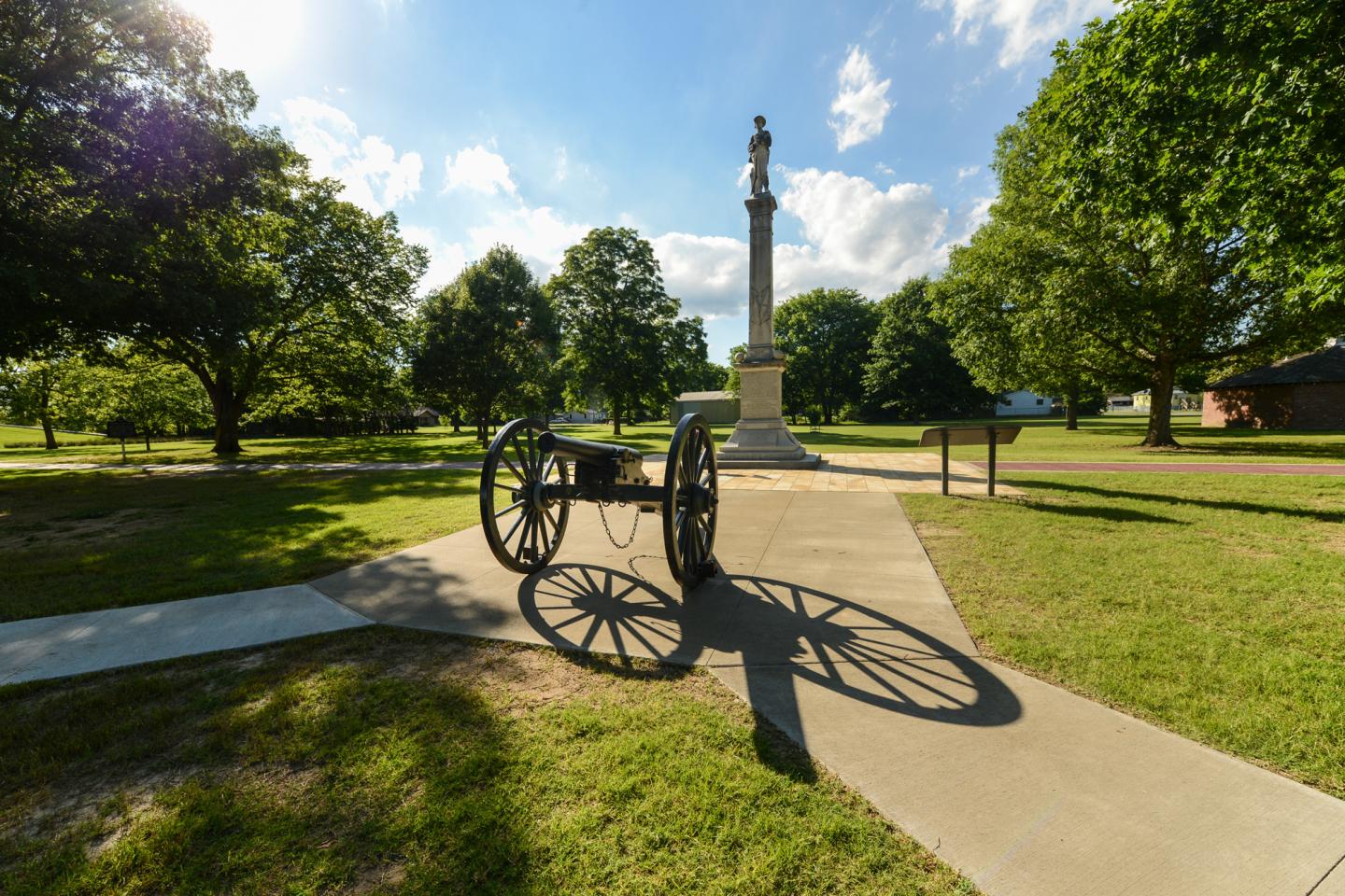 Cannon on a path in a park with trees and a monument in the background.