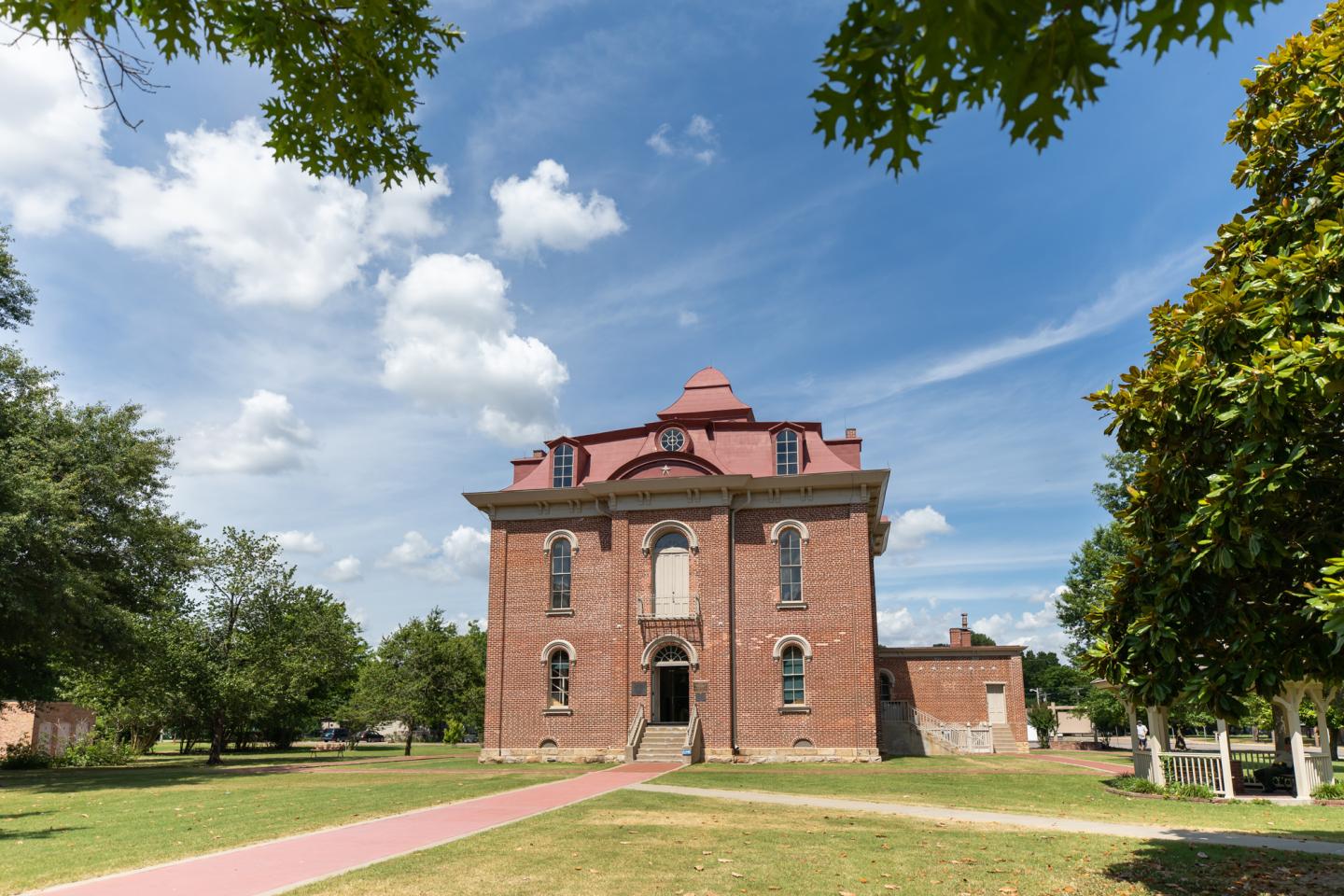 Historic brick building under a blue sky with clouds, surrounded by green trees.