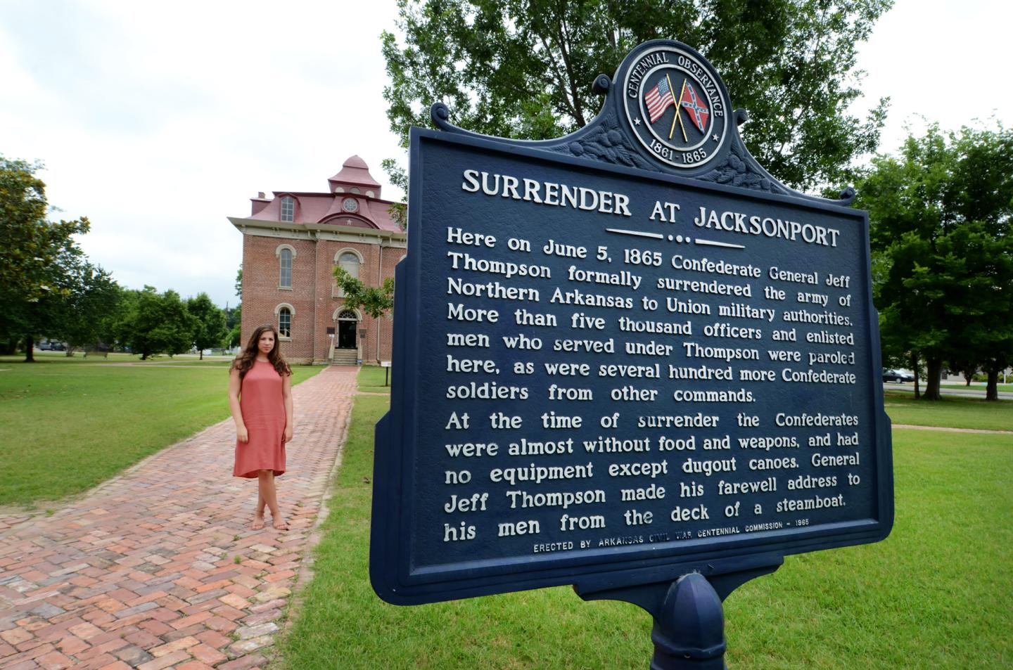 Historic marker with text, woman in red dress, brick path, tree-lined lawn, building in background.