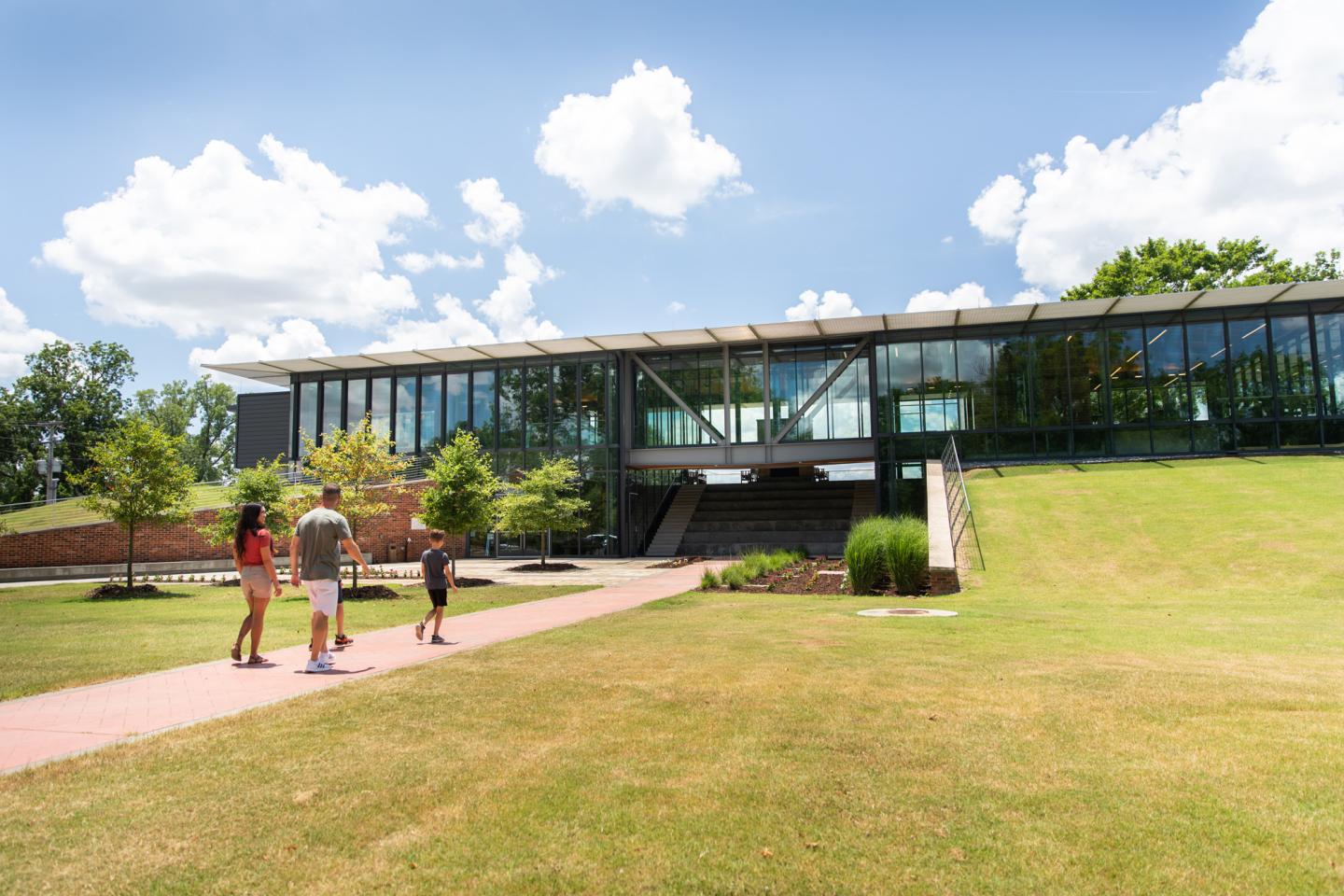 Family walking toward modern glass building under a blue sky with clouds.