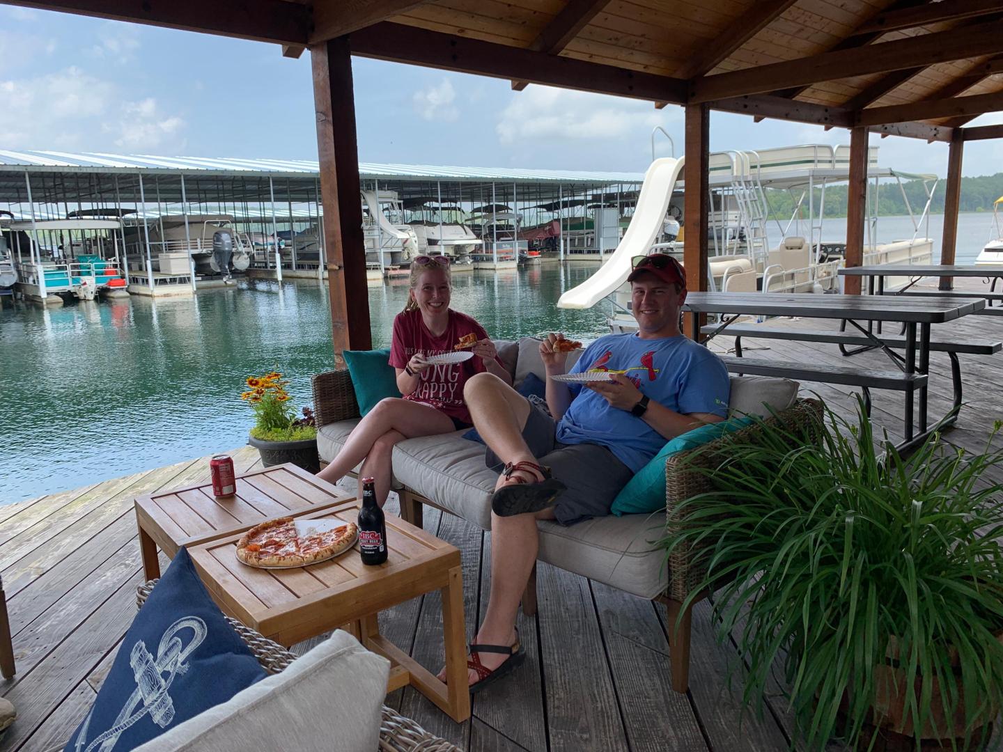 Two people sit on a patio by a marina, eating pizza and relaxing.