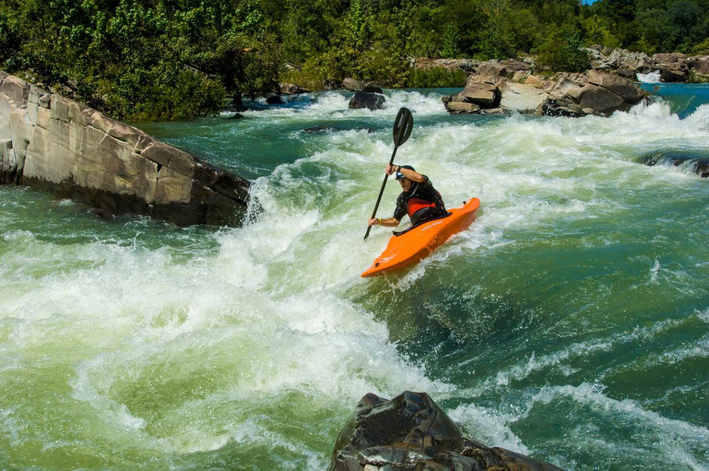 Kayaker navigating rapids in an orange kayak under bright sunlight.