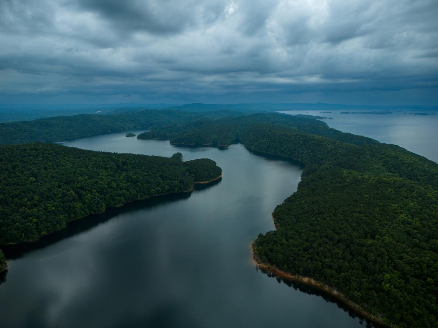 Aerial view of a serene lake surrounded by lush, forested land under a cloudy sky.