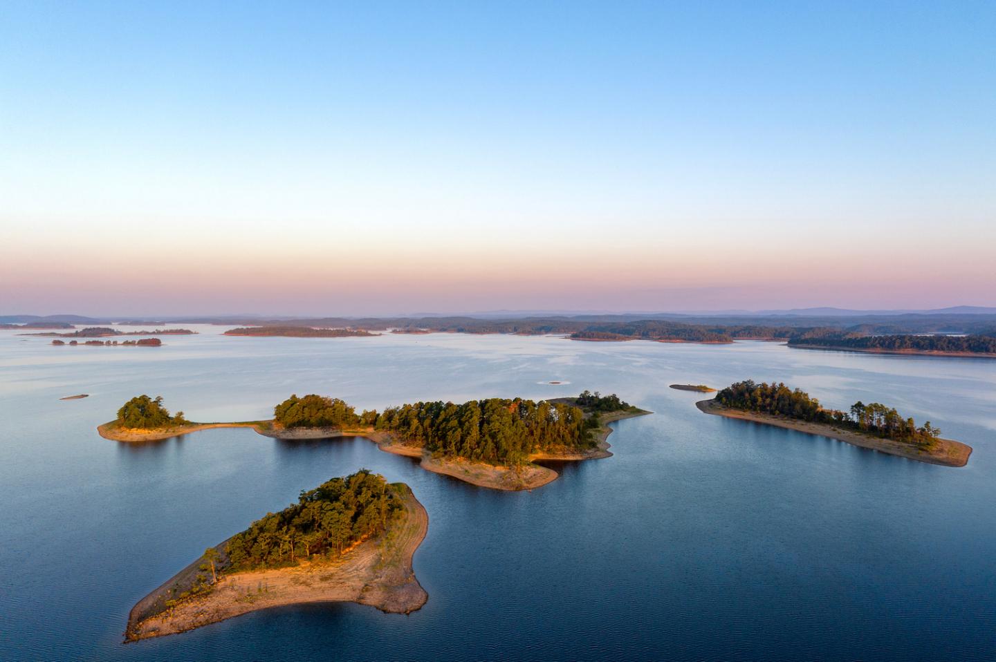 Calm lake with small green islands under a clear blue sky at sunset.