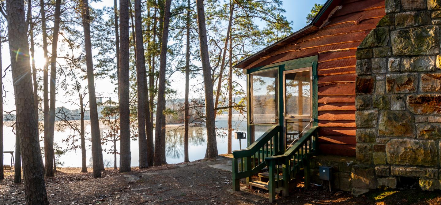 Cabin by a lake, surrounded by pine trees, with sunlight filtering through.