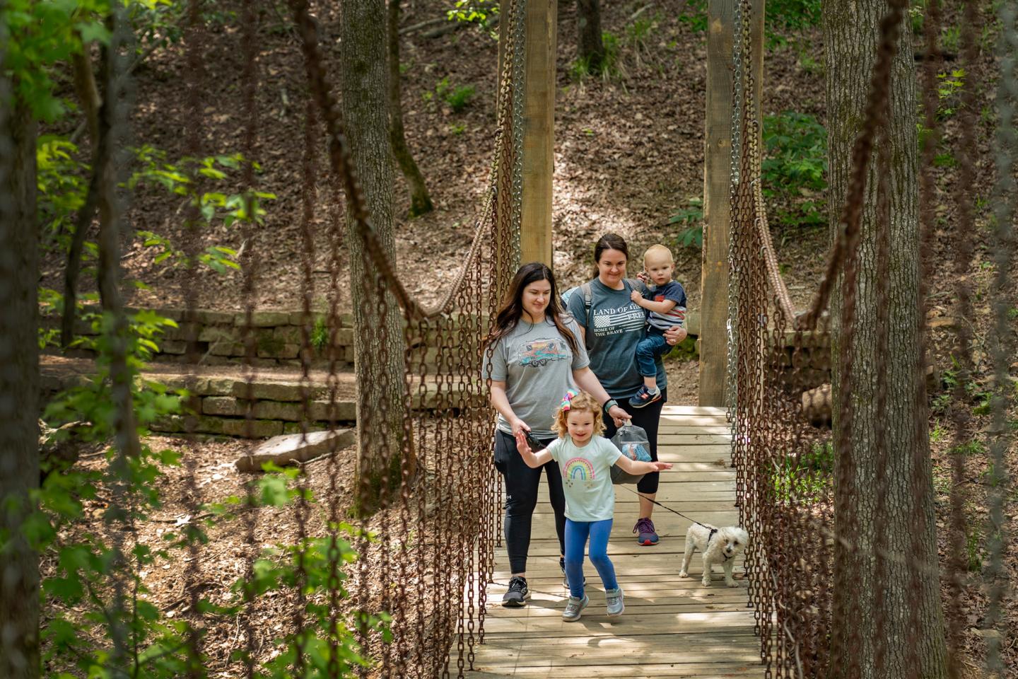 Family walking on a wooded suspension bridge, smiling and holding hands.