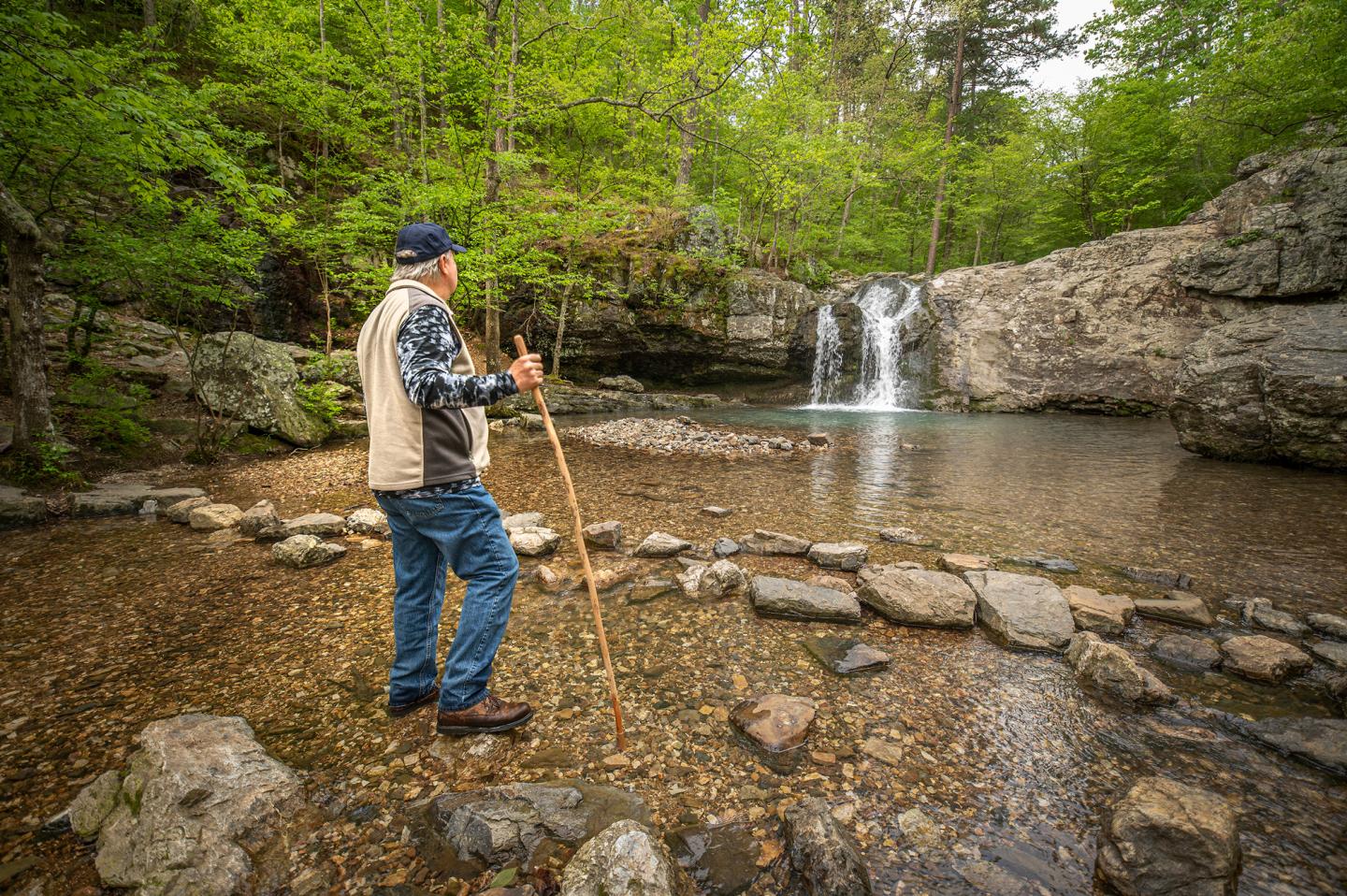 Man with a walking stick by a rocky stream and waterfall.