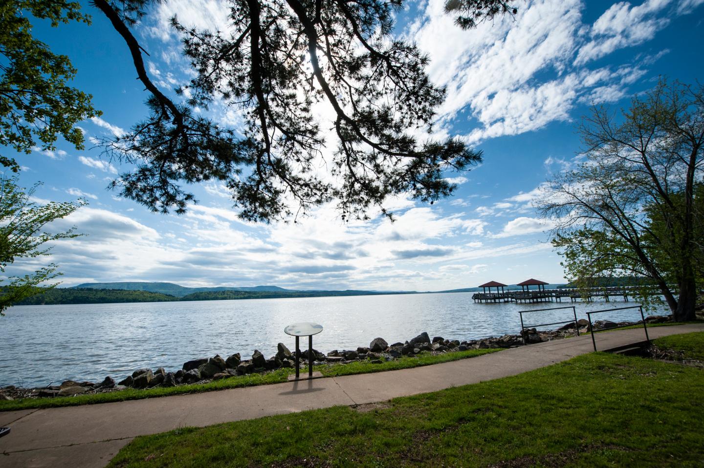 Pathway by a lake under a blue sky with clouds, framed by trees.