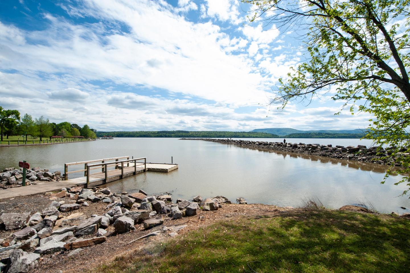 Lake view with a small pier, surrounded by rocks and trees under a blue sky.