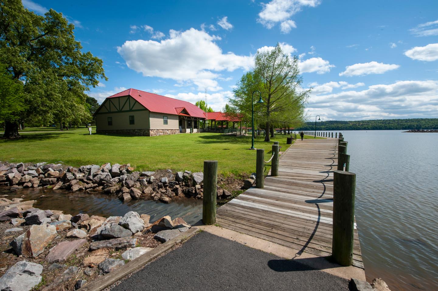 Pathway and dock by a lake, with a red-roofed building, green grass, and a blue sky.