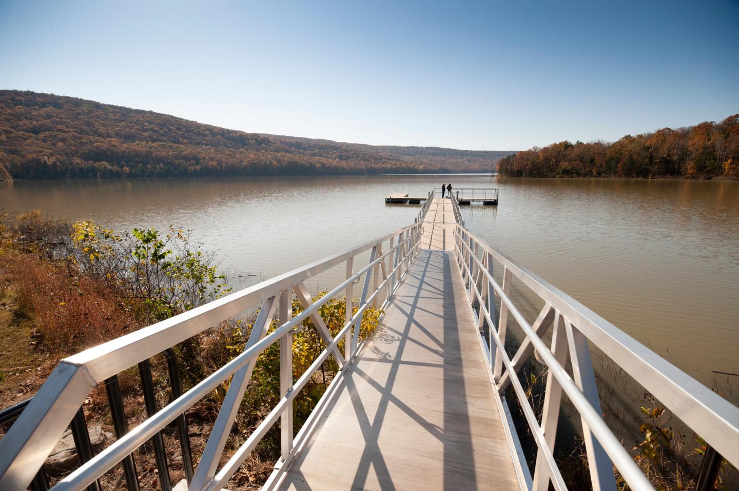 Dock with railing leading to a calm lake surrounded by hills under a clear blue sky.