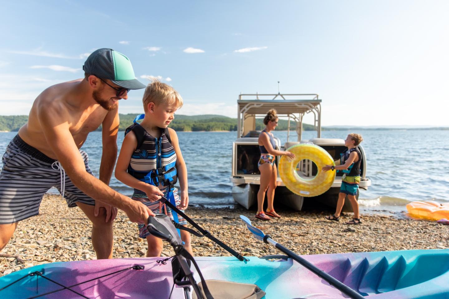 Family preparing kayaks on a sunny lakeside.