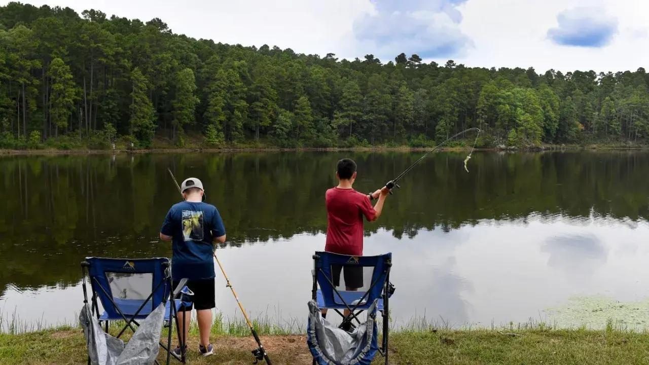 Two people fishing by a forested lake, with folding chairs on the grass.
