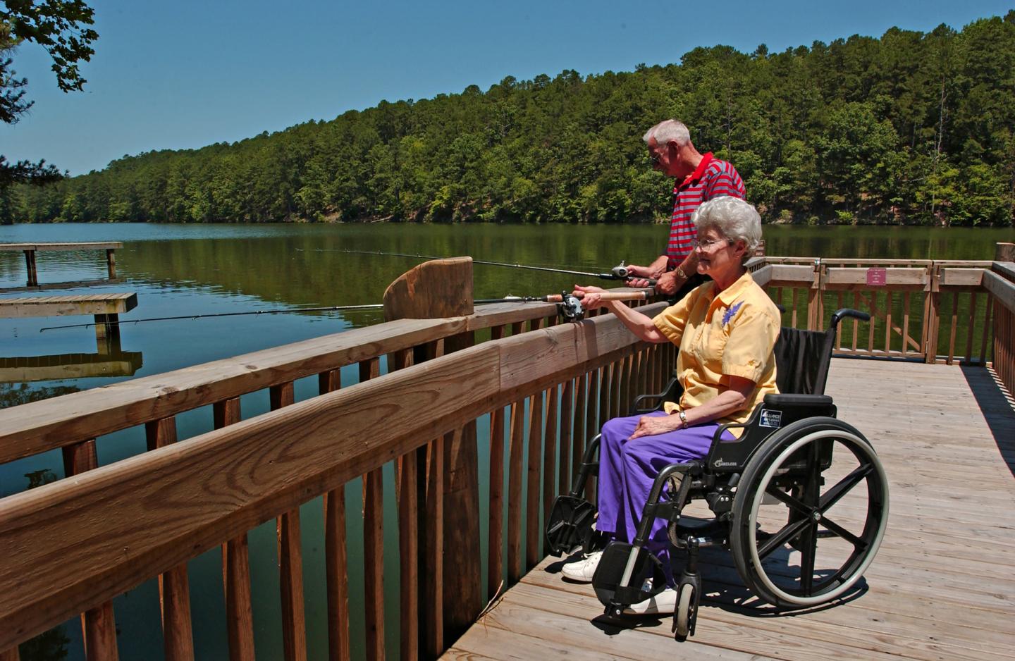 Two elderly people fishing on a wooden dock by a lake, surrounded by trees.