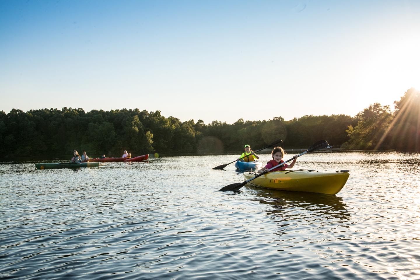 Kayakers paddling on a lake at sunset with a tree-lined shore.
