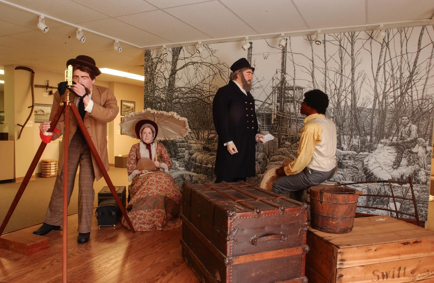 Museum display with historical figures, trunks, and landscape backdrop.