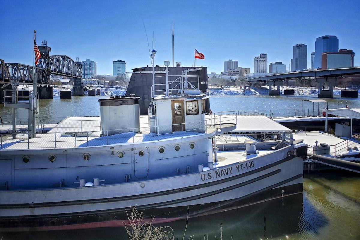 Historic navy tugboat on a river, city skyline in the background.
