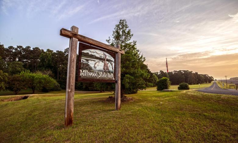 Wooden sign in a grassy park at sunrise, trees and road in the background.