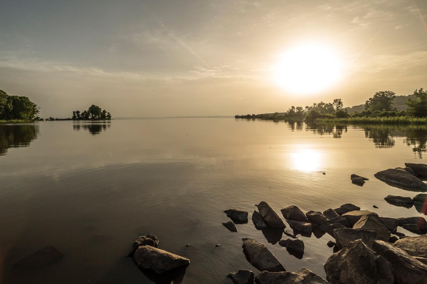 Sunset over a calm lake with scattered rocks in the foreground.