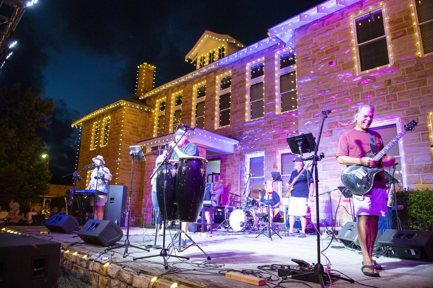 Band performing on outdoor stage with colorful lights at night.