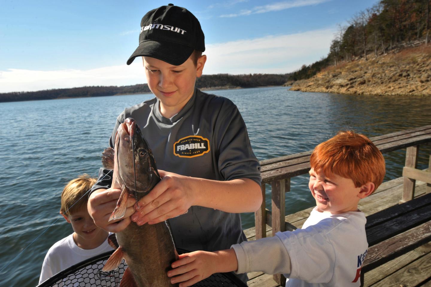 Three boys on a dock, one holding a fish, under a clear blue sky.