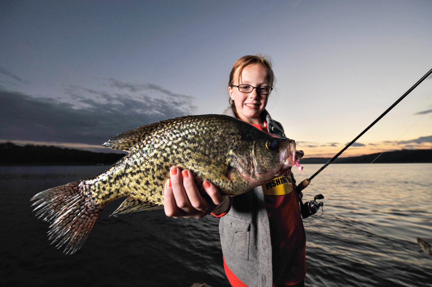 Person holding a fish by a lake at sunset, smiling.