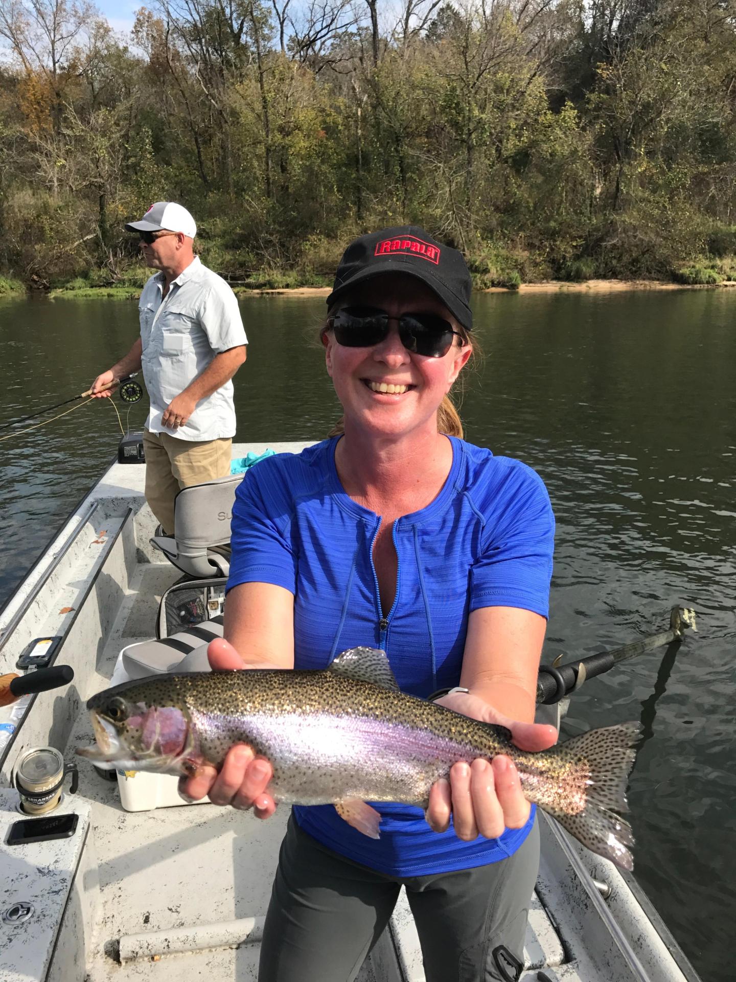Woman smiling on a boat holding a rainbow trout, with a man in the background.