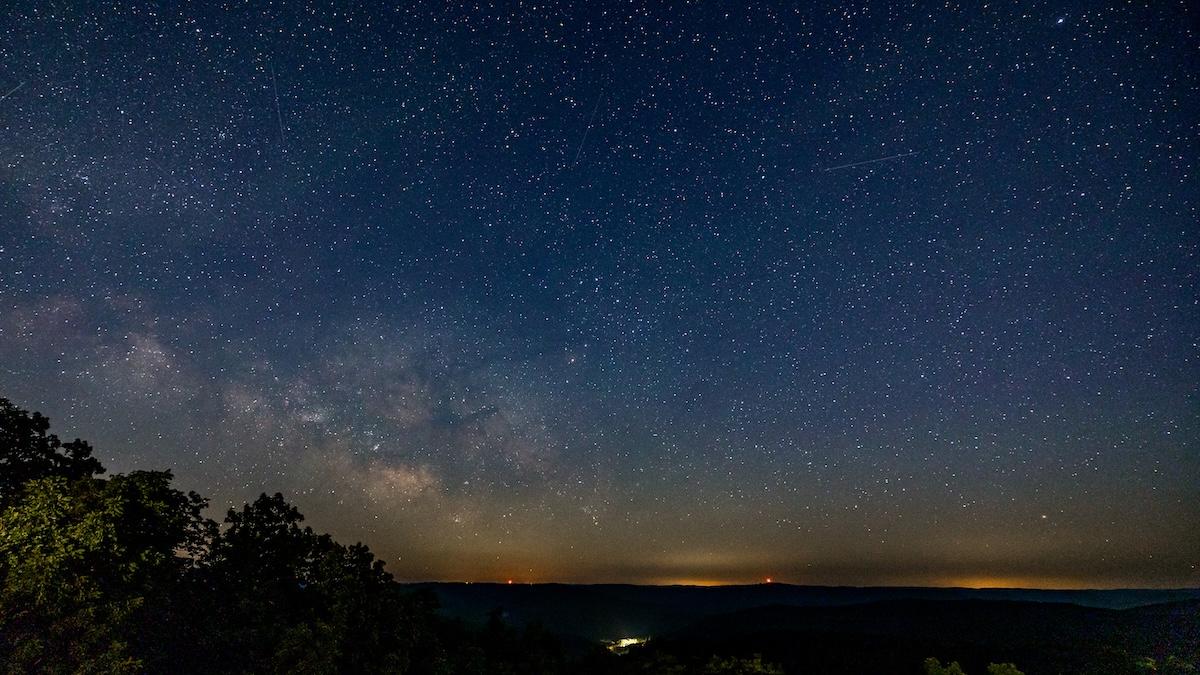 Starry night sky over a dark landscape with faint distant lights.