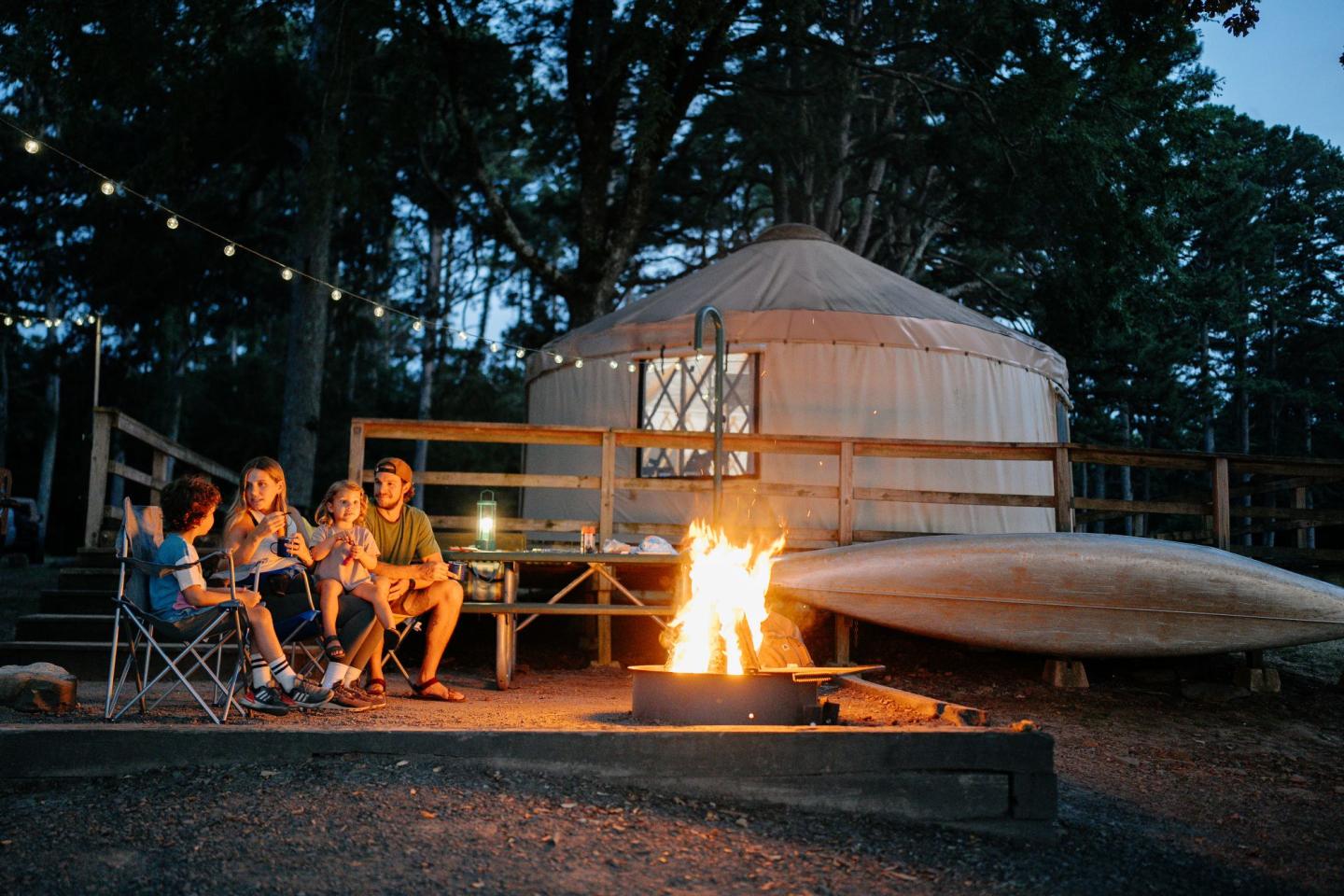Four people sitting around a campfire near a yurt at dusk.